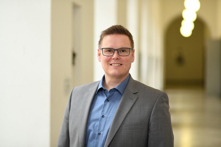 Prof. Rausch hat kurze blonde Haare und eine Brille. Er trägt ein graues Jacket und ein blaues Hemd.